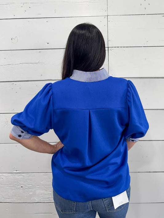 A woman viewed from behind wearing a bright royal blue blouse with puff sleeves and a striped blue-and-white collar, paired with blue jeans, standing against a white wood plank wall.