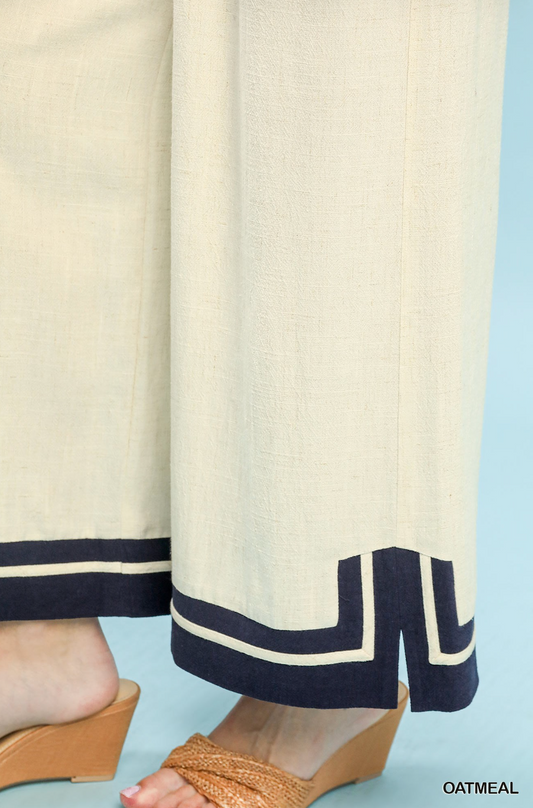Close-up of a person wearing wide-leg cream pants with dark navy geometric trim at the hems, paired with tan wedge sandals against a light blue background.