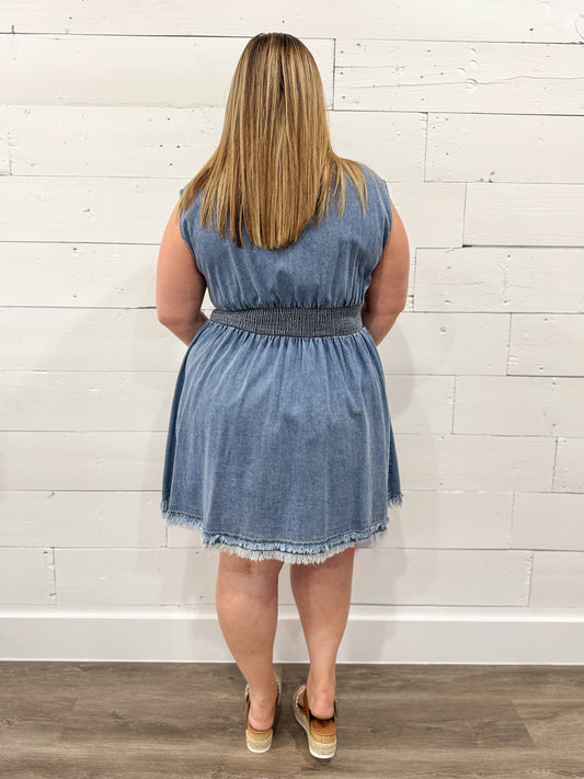 Woman wearing a denim dress standing against a white wooden paneled wall.