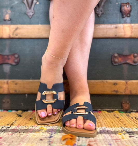 Navy sandals with gold buckles worn by a person on a textured surface with vintage suitcases in the background.