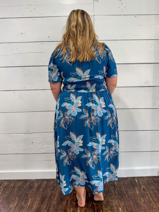 A woman viewed from behind wearing a blue midi dress with a light blue, white, and orange floral pattern, standing against a white wood-paneled wall on a dark wood floor.