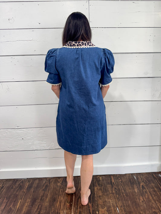 A woman viewed from behind wearing a blue denim-like puff-sleeve dress with a contrasting leopard-print collar, standing against a white wood-paneled wall on a dark wood floor.