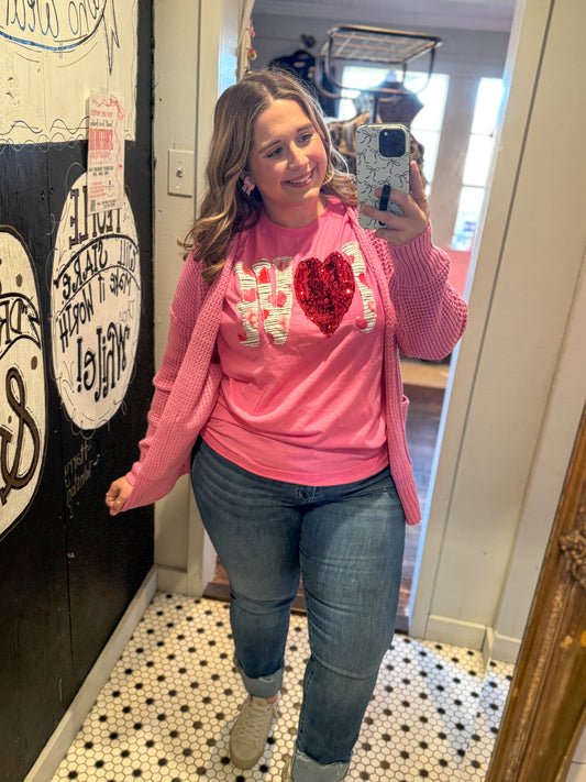 Woman taking a mirror selfie wearing a pink sweater with a red heart design and blue jeans.