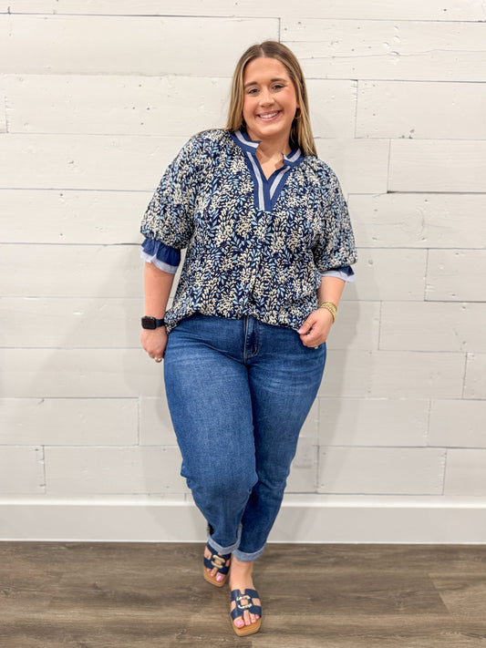 Woman wearing a blue floral blouse and jeans against a white brick wall.
