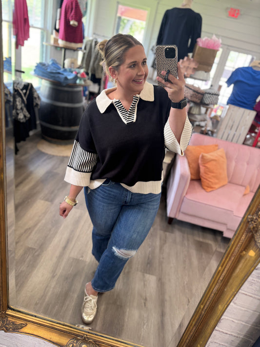 A woman takes a mirror selfie in a boutique while wearing a black knit sweater with cream and white striped details on the collar and sleeves, paired with blue jeans and gold sneakers.