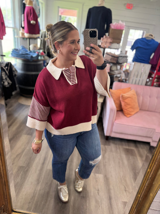 A woman takes a mirror selfie in a clothing boutique wearing a burgundy and cream knit sweater with striped details, blue jeans, and silver sneakers.