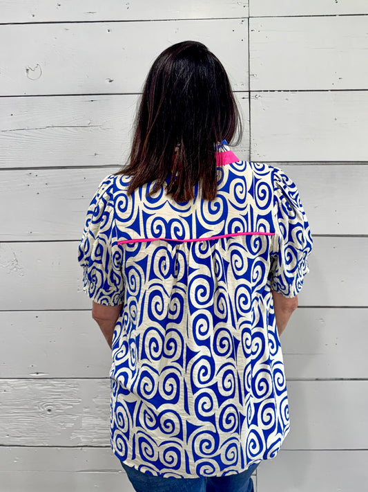 A woman viewed from behind wearing a short-sleeve button-up blouse with a bold blue and white spiral pattern and bright pink trim, standing against a white wood plank wall.