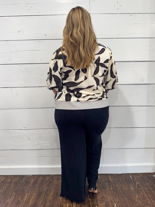 A woman viewed from behind wearing a cream-colored long-sleeve top with a black abstract leaf pattern and a striped hem, paired with dark navy wide-leg pants, standing against a white wood-paneled wall.