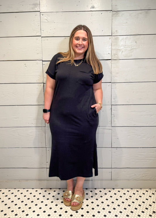 Woman wearing a black dress standing against a light wooden paneled wall.