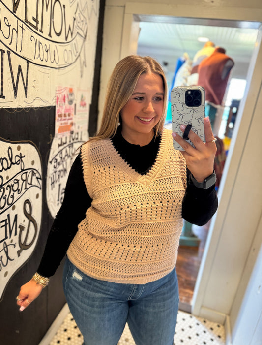 Woman taking a mirror selfie wearing a beige crochet vest and blue jeans.