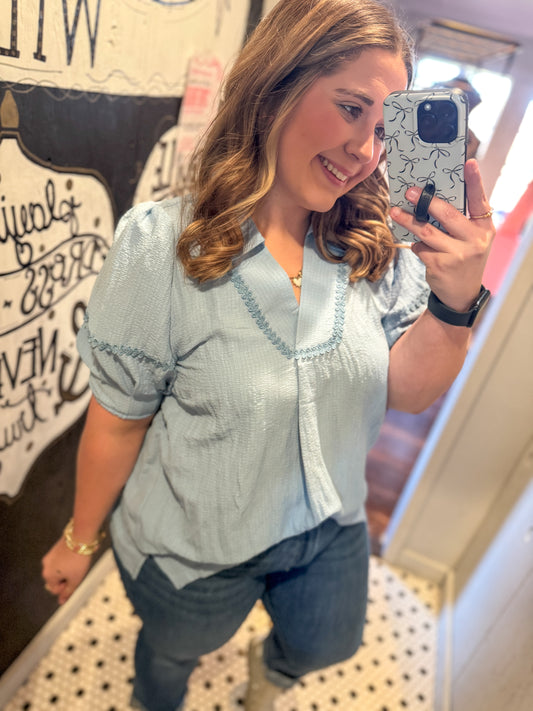 Woman taking a mirror selfie wearing a light blue blouse and jeans.