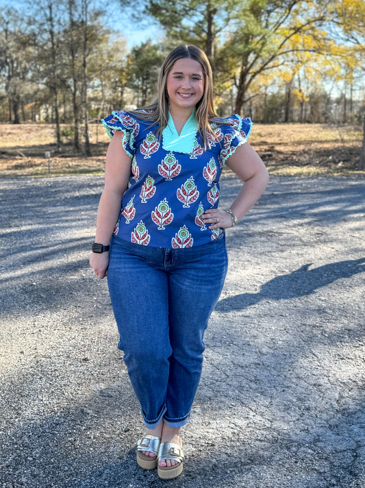 Woman wearing a blue patterned top and jeans standing outdoors on a gravel surface.