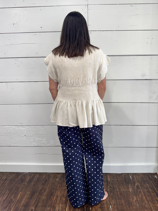 A woman viewed from behind wearing a cream-colored textured peplum top and navy blue wide-leg pants with white polka dots, standing against a white wood-paneled wall on a dark wood floor.
