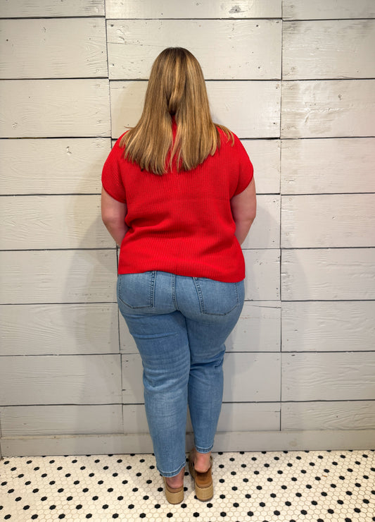 Person wearing a red shirt and blue jeans standing against a light wooden paneled wall.