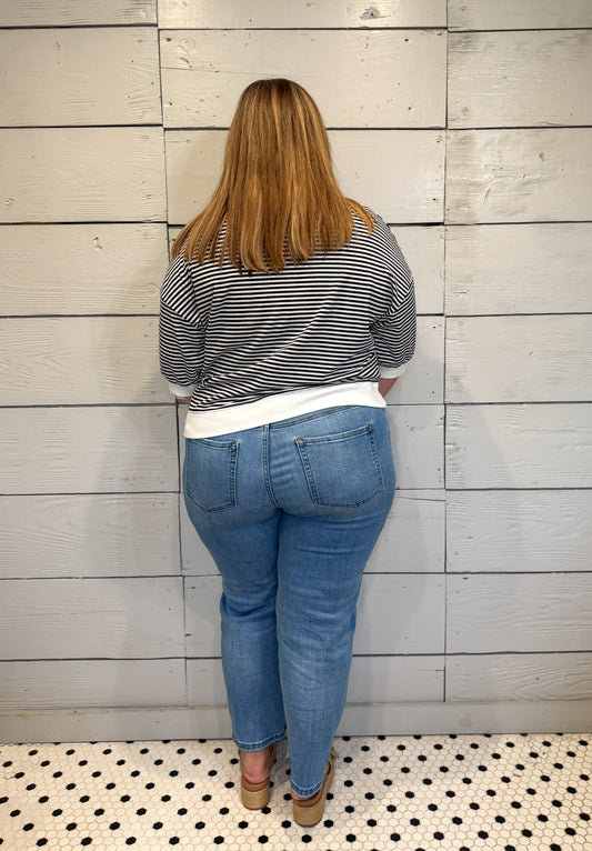 Person wearing a striped shirt and blue jeans standing against a wooden paneled wall.