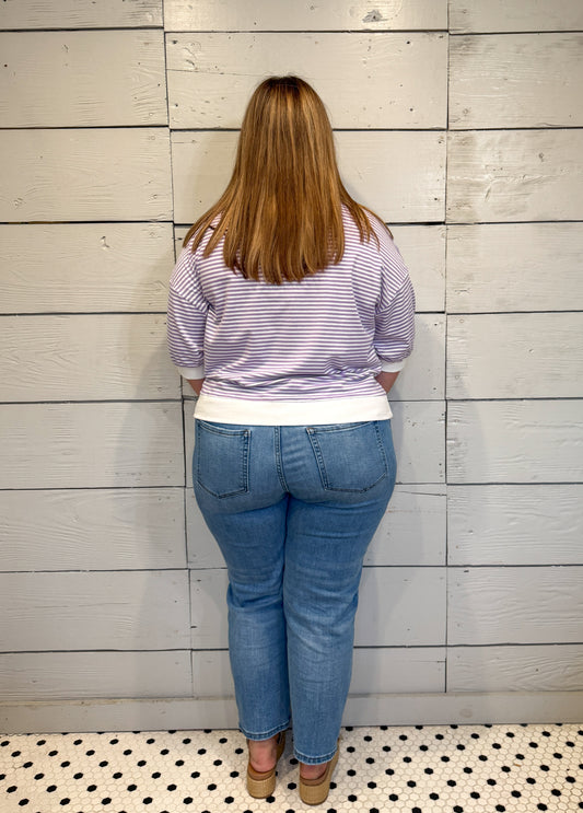 Person wearing a striped shirt and blue jeans standing against a wooden paneled wall.