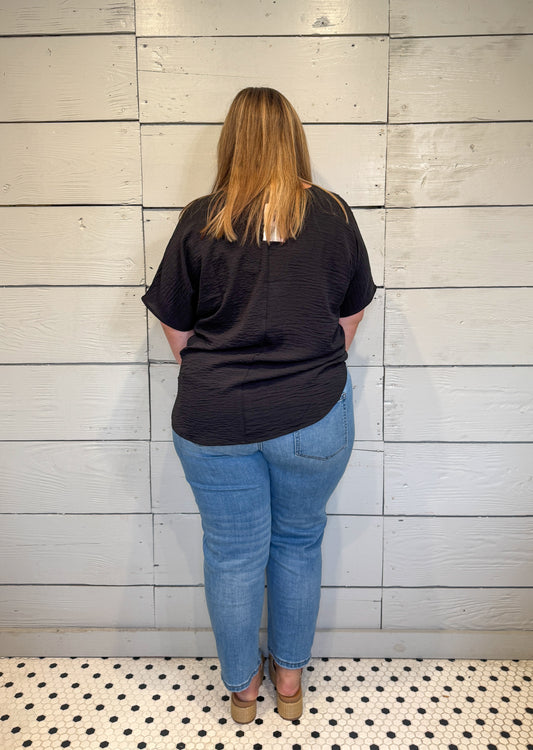 Person wearing a black t-shirt and blue jeans standing against a light wooden panel wall.