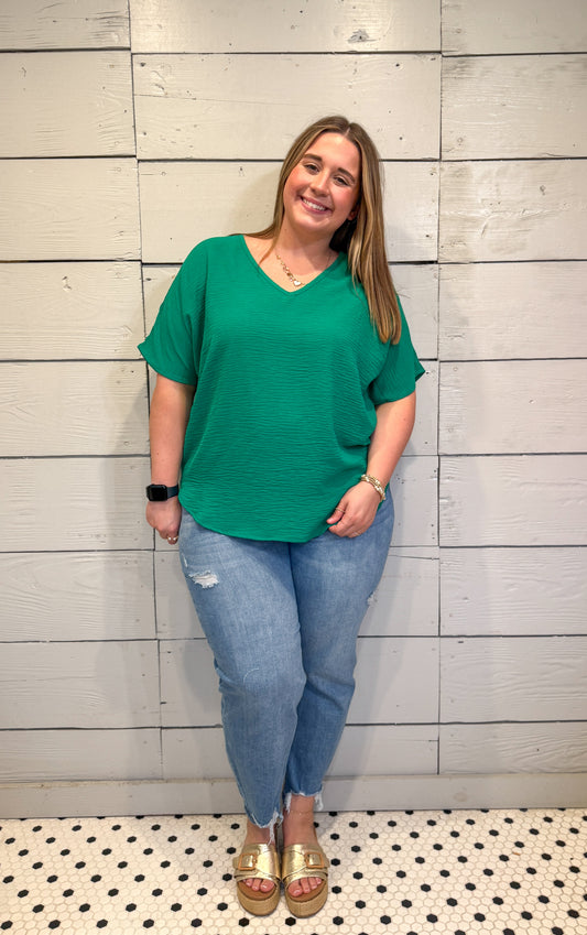 Woman wearing a green top and blue jeans standing against a light wooden paneled wall.