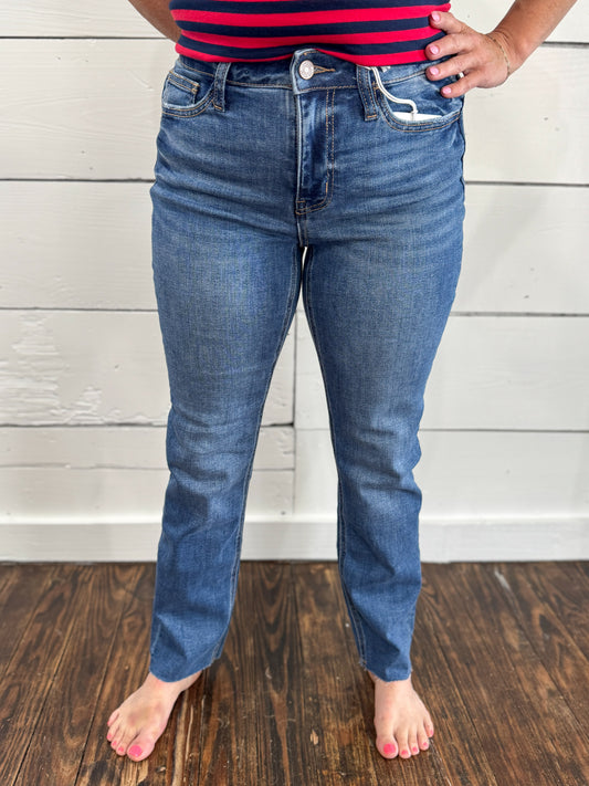 A person wearing medium-wash blue jeans, shown from the waist down, standing barefoot on a dark wood floor against a white wood-paneled wall.