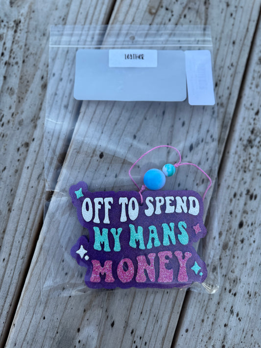 Decorative sign with text 'Off to spend my man's money' on a wooden surface.