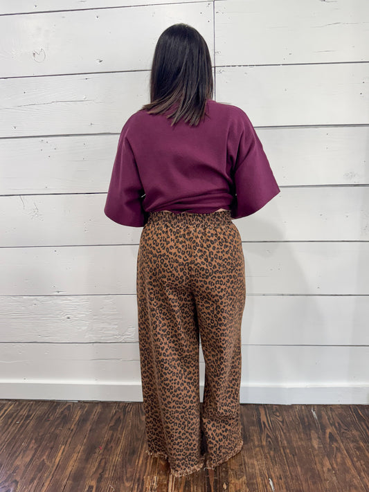 A person standing with their back to the camera, wearing a short-sleeve burgundy top and brown leopard-print pants, set against a white wood-paneled wall and dark wood floor.