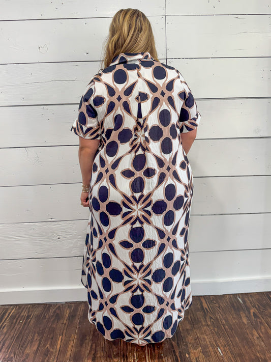 A woman viewed from behind wearing a short-sleeve collared dress with a geometric pattern in navy blue, beige, and white, standing against a white wood-paneled wall.