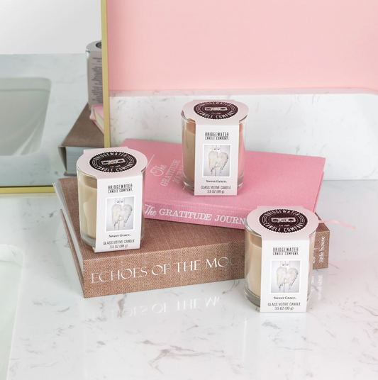 Three glass jar candles with white labels and gold wax tops are arranged on stacked books on a white marble surface, with a pink wall and mirror in the background.