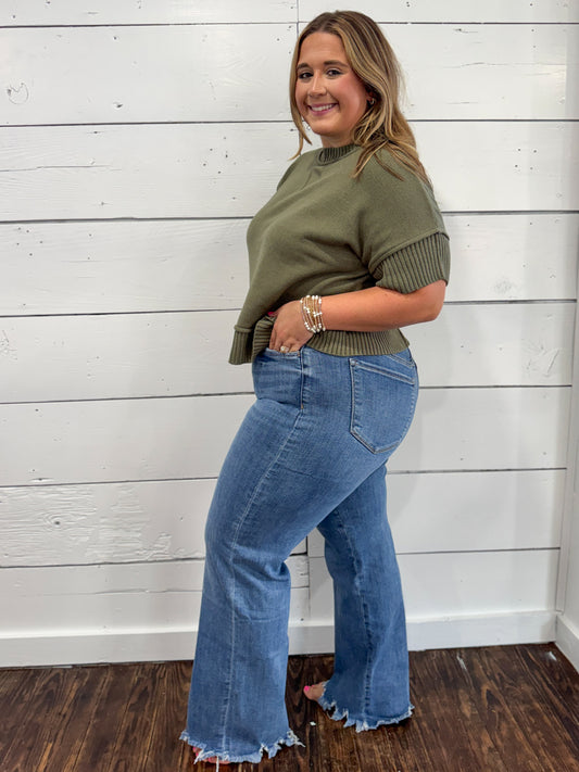 A woman modeling an olive green mock neck top with ribbed short sleeves and a ribbed hem, paired with blue flare jeans, standing against a white wood-paneled wall.