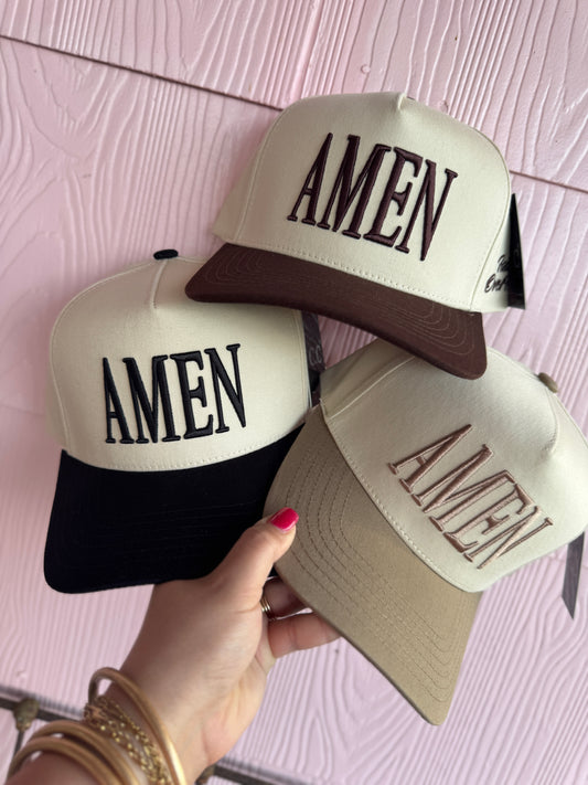 Three 'AMEN' caps held by a hand against a pink textured background