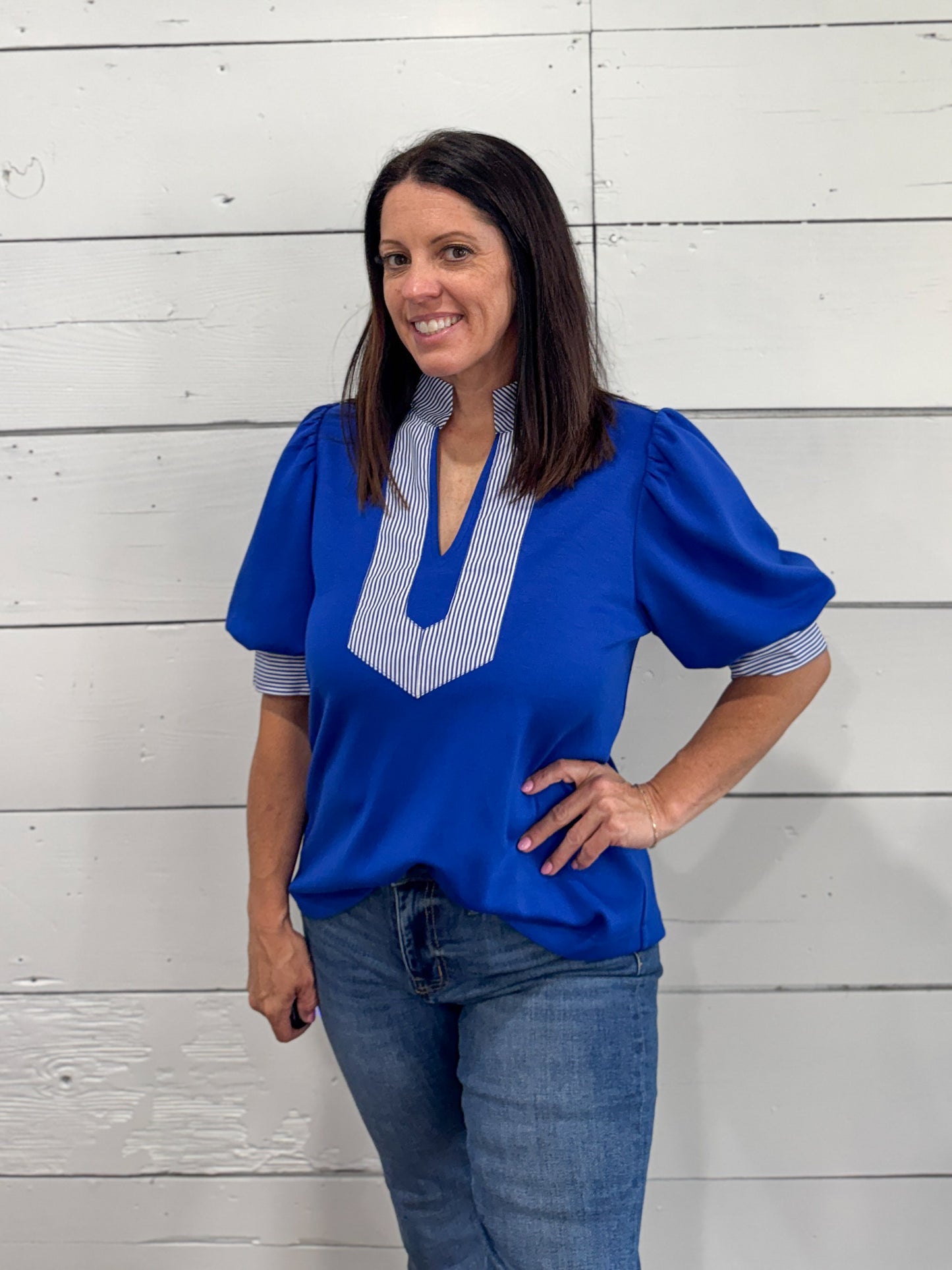 A woman wearing a royal blue short-sleeve blouse with a striped V-neck panel and blue jeans, standing against a white wood plank wall.