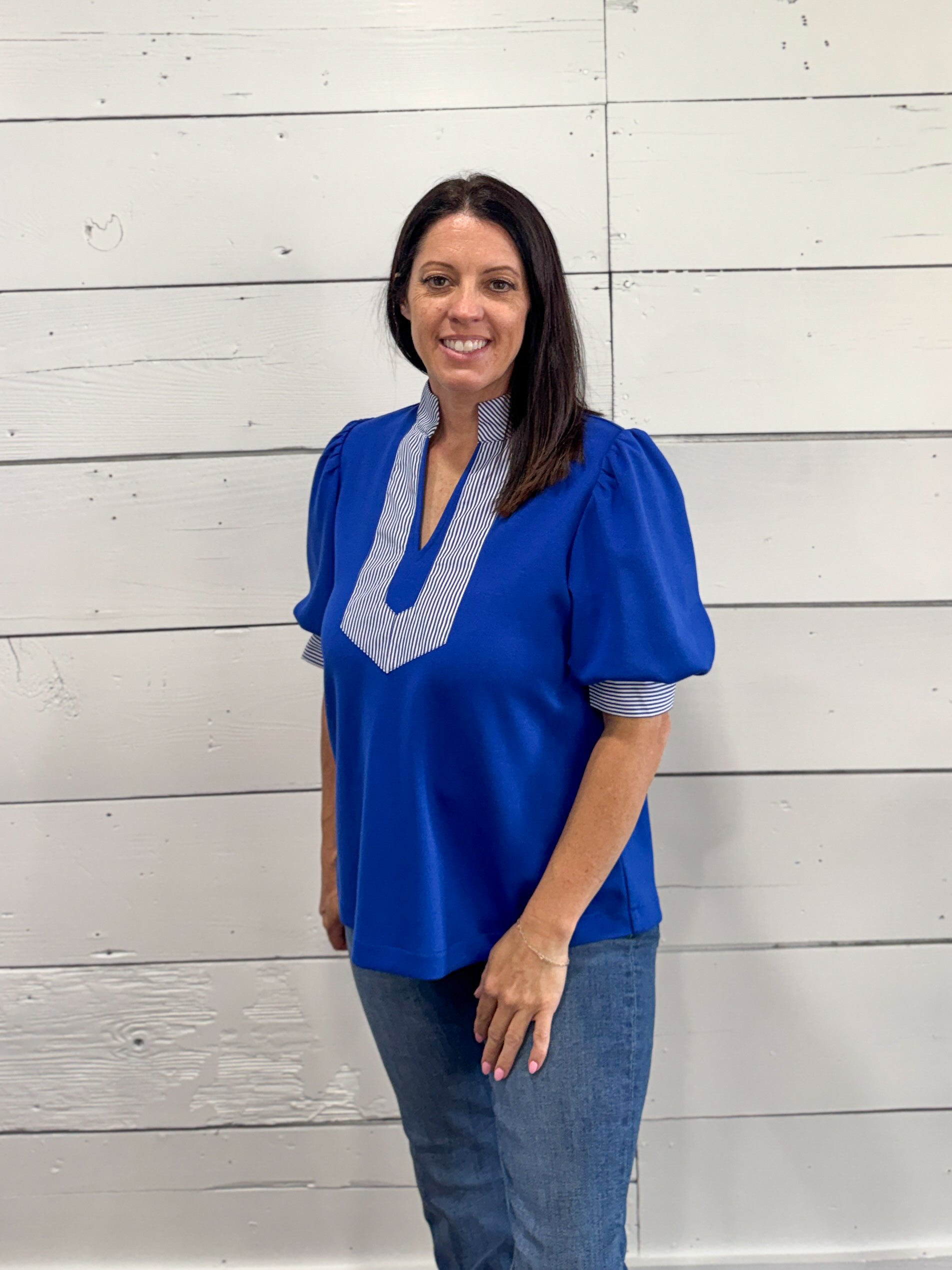 A woman wearing a royal blue short-sleeve blouse with a light blue and white striped V-neck trim, paired with blue jeans, standing against a white wood plank wall.