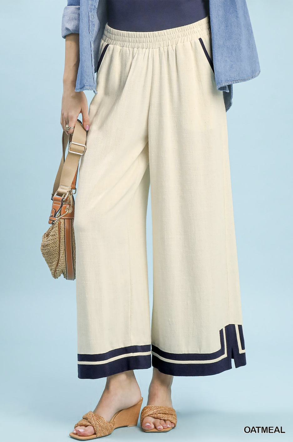 A person wearing wide-leg cropped pants in a warm off-white/cream color with navy and cream stripes at the hem, styled with a light blue denim shirt, a navy top, and tan wedge sandals. The model is holding a woven straw bag against a solid light blue background.