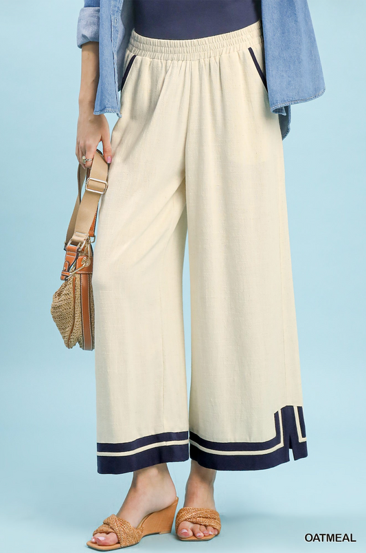 A person wearing wide-leg cropped pants in a warm off-white/cream color with navy and cream stripes at the hem, styled with a light blue denim shirt, a navy top, and tan wedge sandals. The model is holding a woven straw bag against a solid light blue background.