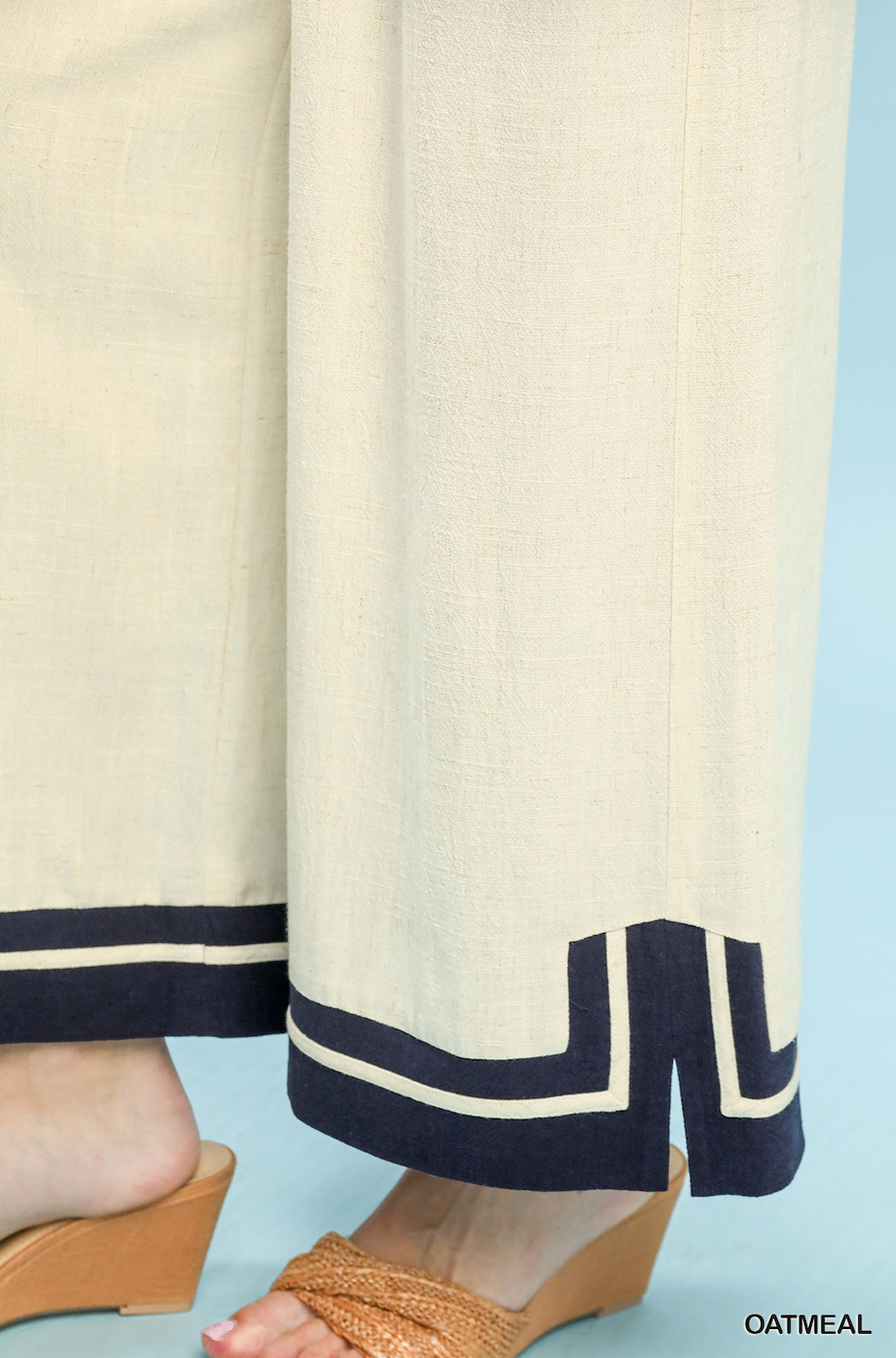 Close-up of a person wearing wide-leg cream pants with dark navy geometric trim at the hems, paired with tan wedge sandals against a light blue background.