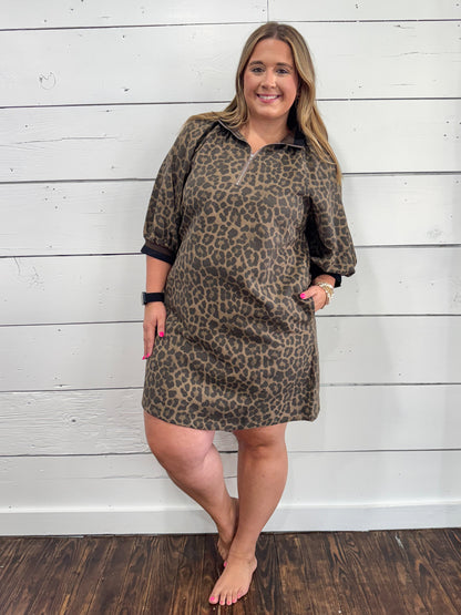 A woman modeling a brown and black leopard print polo shirt dress with three-quarter sleeves, standing against a white wood-paneled wall.
