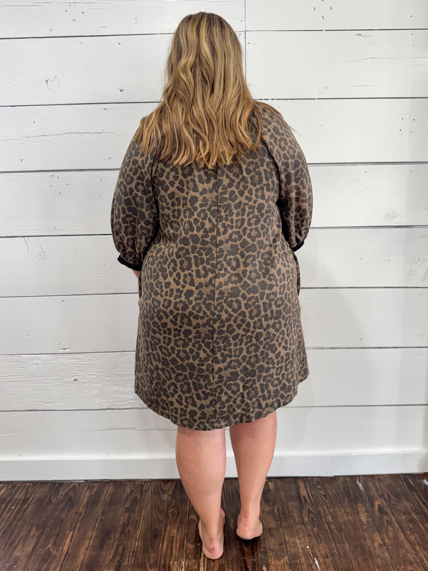 A woman viewed from behind wearing a leopard print dress with long sleeves, standing against a white wood-paneled wall on a dark wood floor.