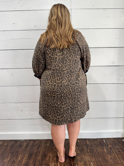 A woman viewed from behind wearing a leopard print dress with long sleeves, standing against a white wood-paneled wall on a dark wood floor.