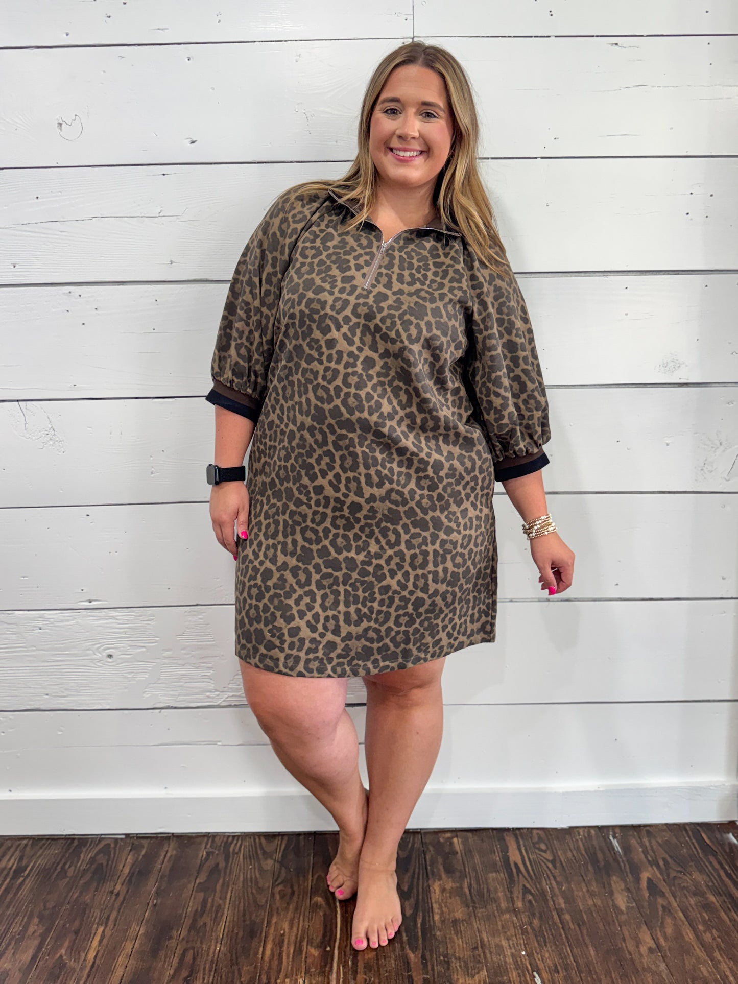 A woman modeling a brown and black leopard print polo shirt dress with three-quarter sleeves and black ribbed cuffs, standing against a white wood-paneled wall.