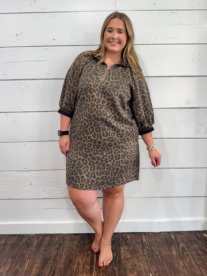 A woman modeling a brown and black leopard print polo shirt dress with three-quarter sleeves and black ribbed cuffs, standing against a white wood-paneled wall.
