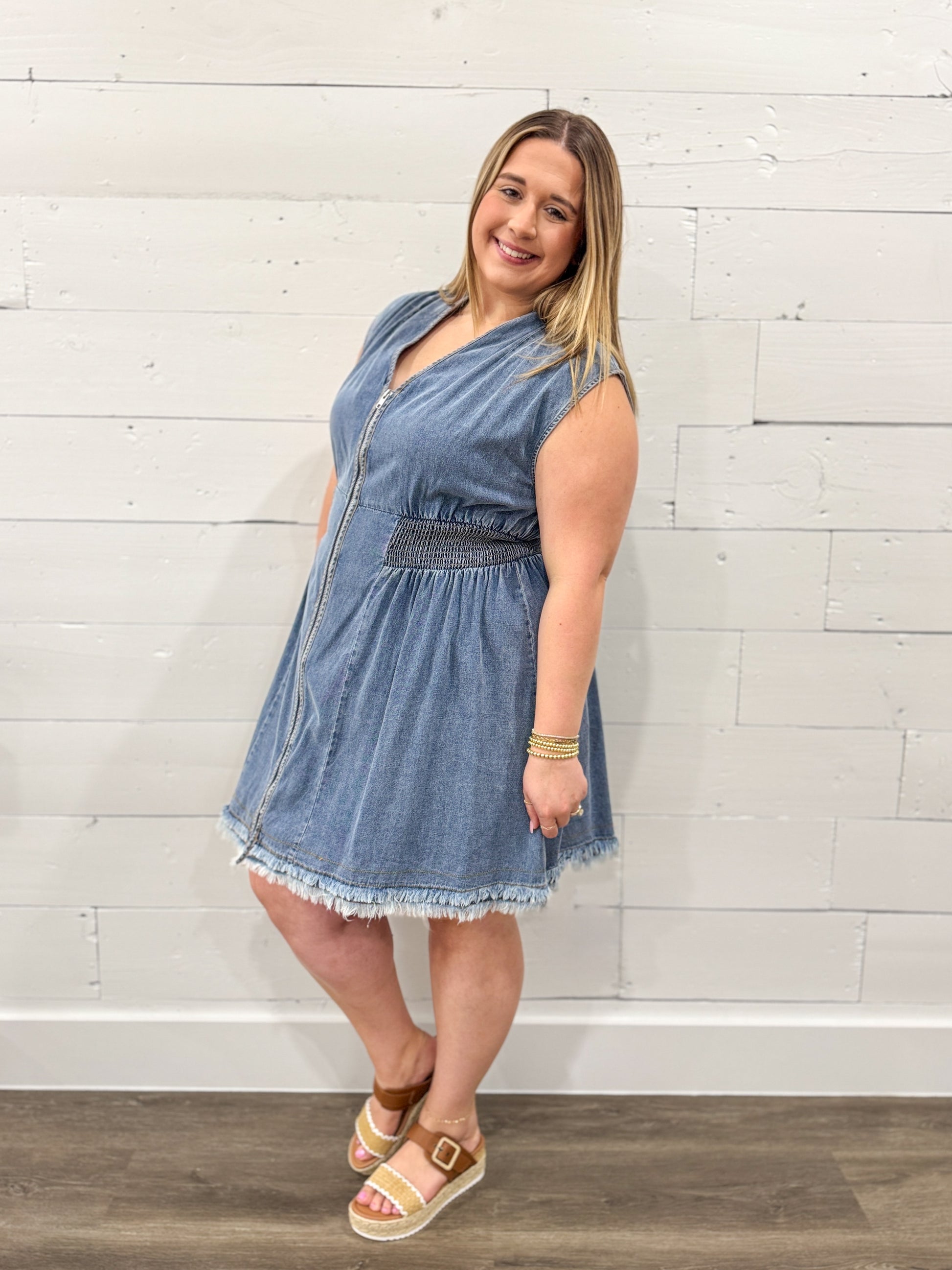 Woman wearing a denim dress standing against a white brick wall.