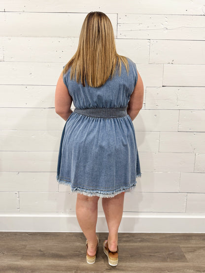 Woman wearing a denim dress standing against a white wooden paneled wall.