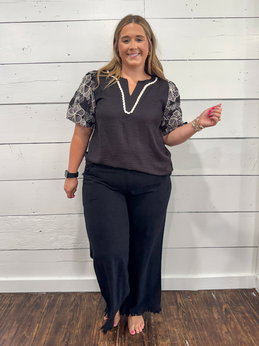 A woman modeling a dark gray V-neck top with patterned short sleeves and black wide-leg pants, standing against a white shiplap wall.