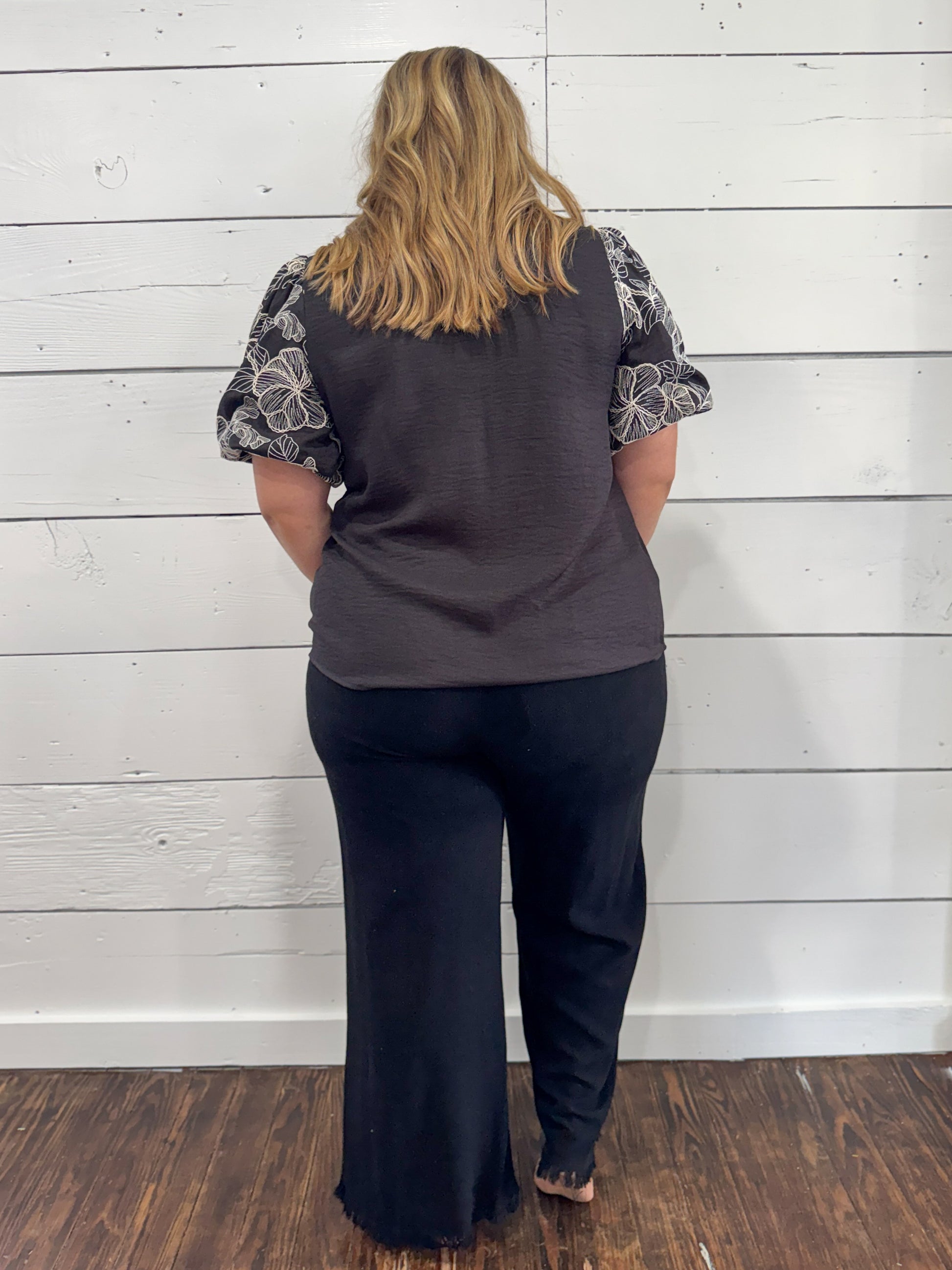 A woman viewed from behind wearing a dark gray sleeveless top with patterned short sleeves and dark wide-leg pants, standing against a white wood plank wall on a dark wood floor.
