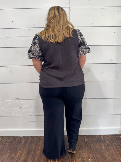 A woman viewed from behind wearing a dark gray sleeveless top with patterned short sleeves and dark wide-leg pants, standing against a white wood plank wall on a dark wood floor.