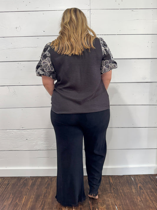A woman viewed from behind wearing a dark gray sleeveless top with patterned short sleeves and dark wide-leg pants, standing against a white wood plank wall on a dark wood floor.