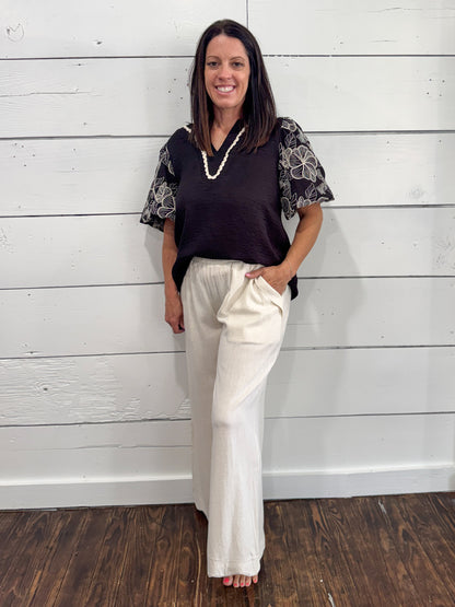 A woman stands against a white wood-paneled wall wearing a dark brown sleeveless top with patterned short sleeves and off-white wide-leg pants.
