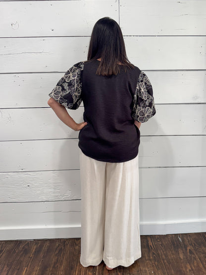 A woman viewed from behind wearing a dark brown sleeveless vest over a black and white patterned short-sleeve top, paired with cream wide-leg pants, standing against a white wood-paneled wall.