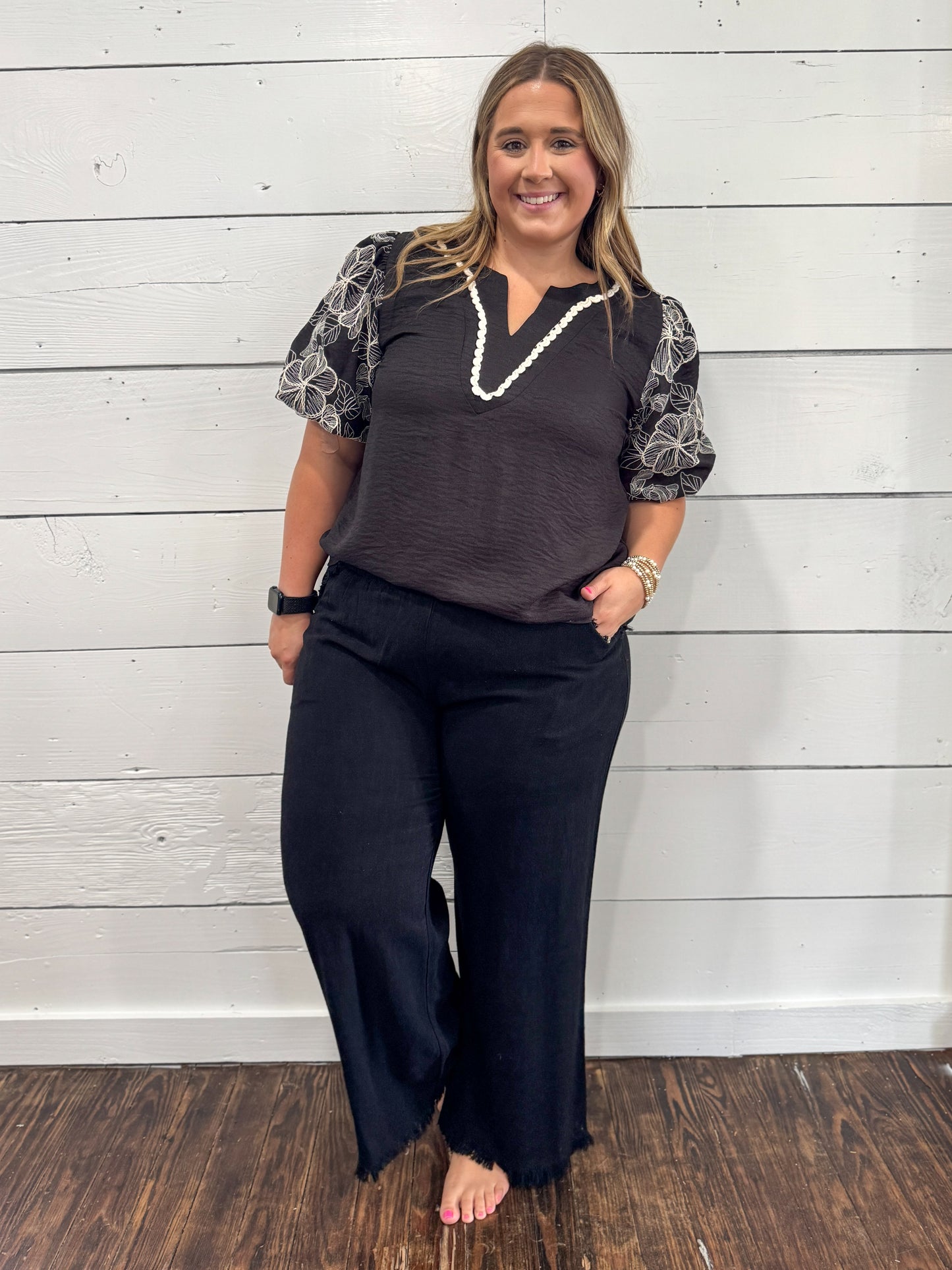 A woman modeling a dark gray V-neck top with patterned short sleeves and black wide-leg pants, standing against a white wood-paneled wall.