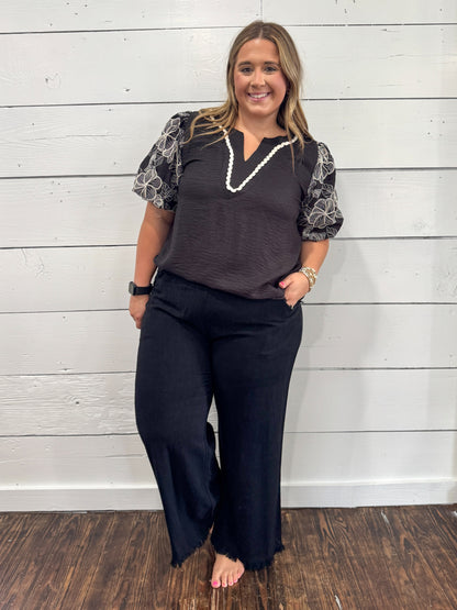A woman modeling a dark gray V-neck top with patterned short sleeves and black wide-leg pants, standing against a white wood-paneled wall.