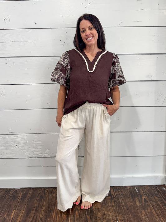 A woman stands barefoot against a white wood-paneled wall wearing a brown sleeveless vest top with sheer patterned sleeves and cream wide-leg pants.